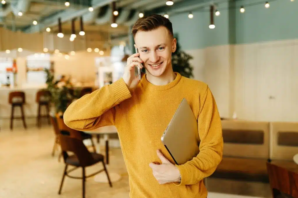 Young guy in yellow jacket uses technology; freelance; student; man; work; laptop; smile; boy; communicate; connect; cafe; inside; successful; workplace; restaurant; cup of coffee; lifestyle; mobile phone; portrait; fashion; stylish; casual; bright; person; cool; hipster; laugh; stand up; online; business; businessman; small business; manager; boss; thoughtful; serious; sitting; headphones; wireless technology; typing; watch; busy; indoor; fun; modern; interior; design; public space; male; happy;workspace
